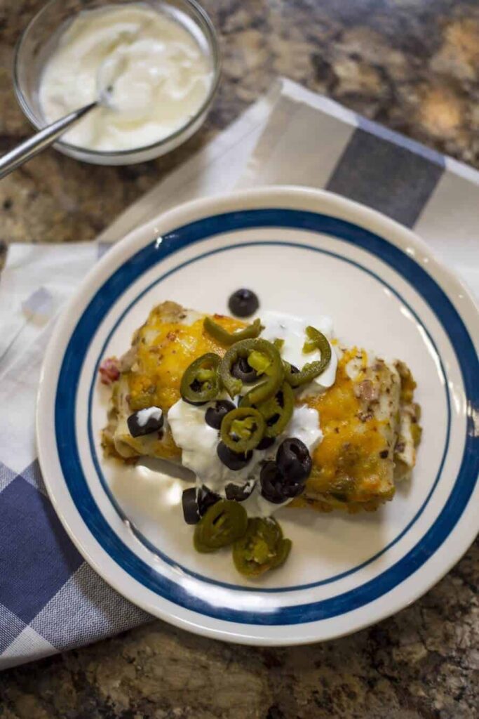 overhead photo of a plate with blue rim. Cheese and green chili covered pork burrito with olives, sour cream and jalapeno pepper.