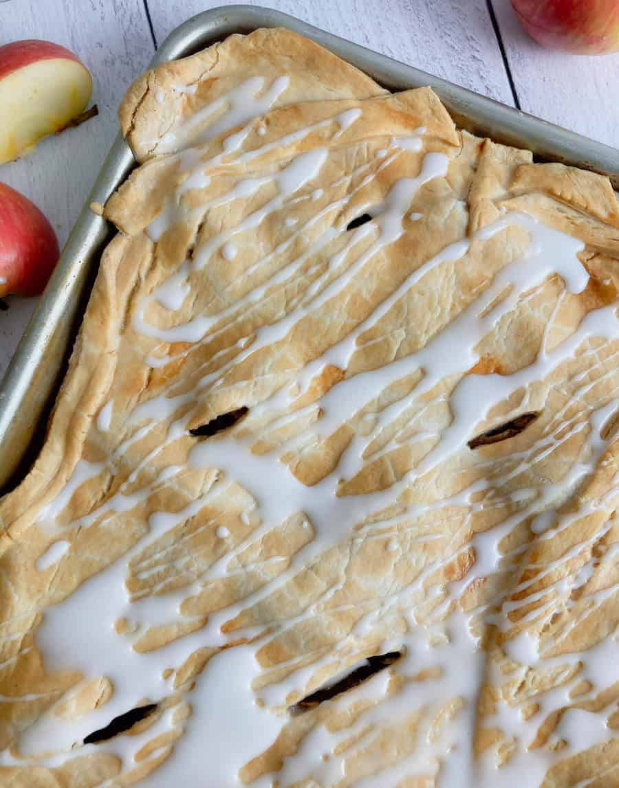 An apple slab pie on the counter surrounded by apples and drizzled with icing.