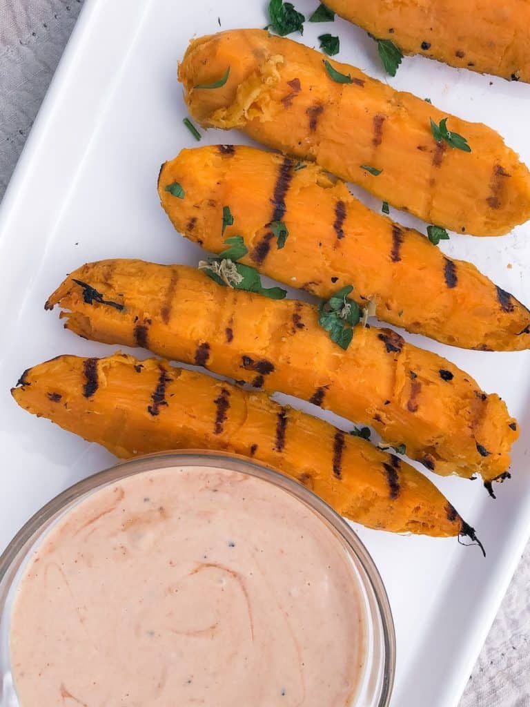 four grilled sweet potato fries with green parsley on a white plate with a bowl of dipping sauce