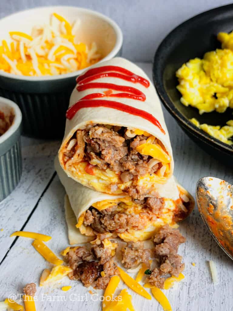 Two rolled breakfast burritos with sausage and eggs spilling out, topped with siracha.