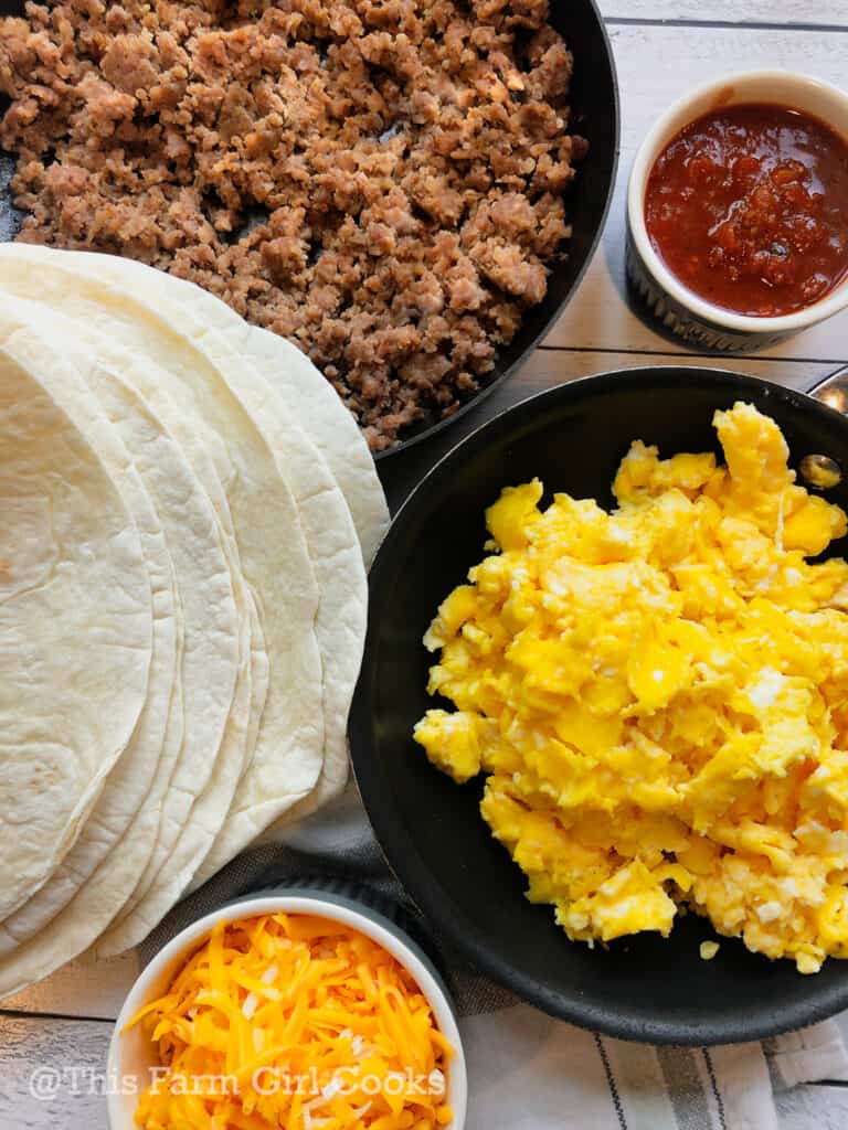 Flour tortillas, shredded cheddar cheese, scrambled eggs, cooked pork sausage and chunky salsa on a table.