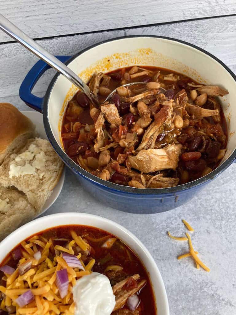 A ladle full of pulled pork chili with rolls on the side.