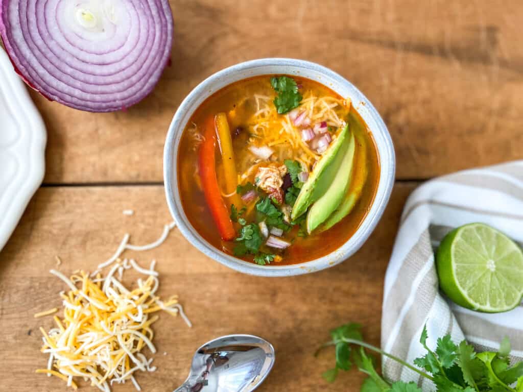 A bowl of chicken fajita soup surrounded by toppings of lime and shredded cheese.