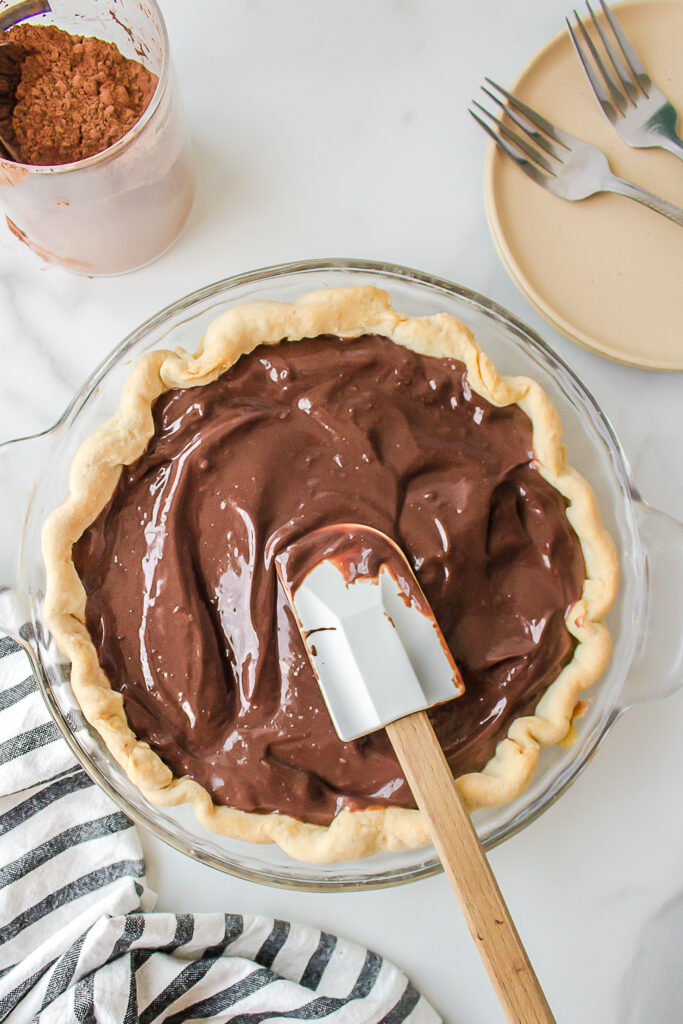 A pie pan and pie crust with chocolate custard mixture being smoothed by a rubbed spatula.