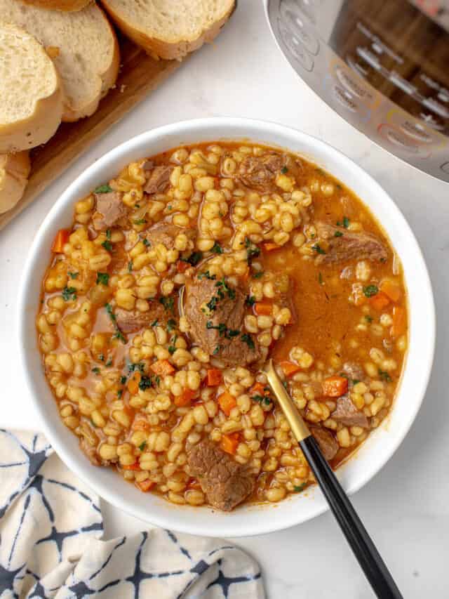 A hearty bowl of barley soup with beef and vegetables, with a spoon in it.