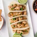 Five street tacos on a rectangle plate garnished with lime halves.
