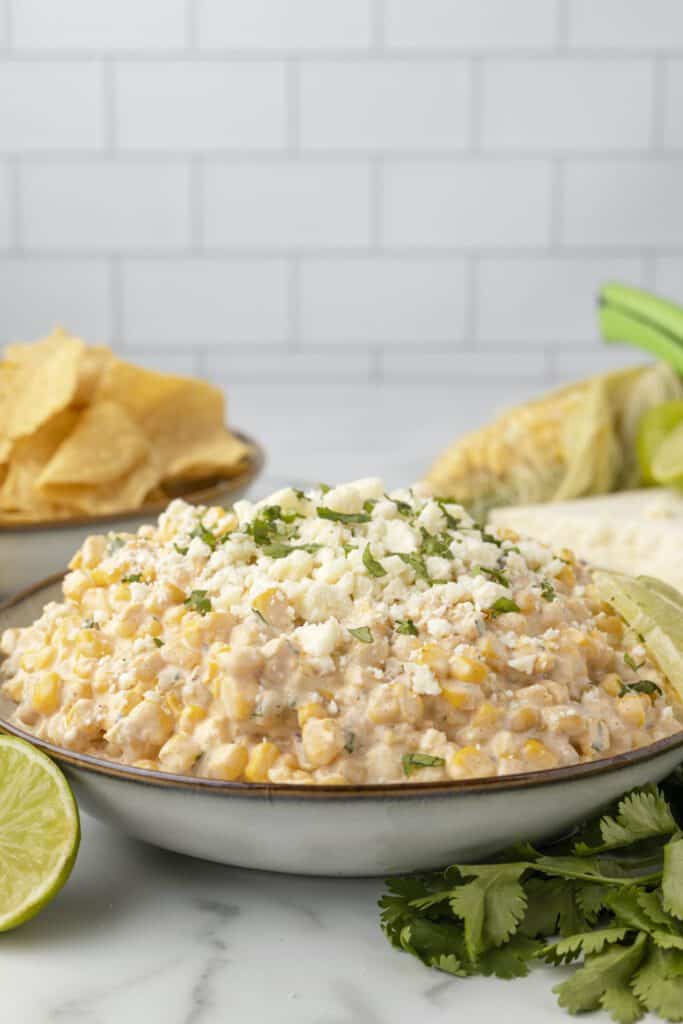 Mexican street corn dip heaped in a bowl surrounded by lime, cilantro and tortilla chips.