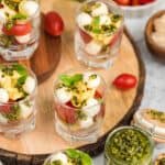 Tortellini Caprese salad cups on a wooden slab surrounded by fresh pesto.