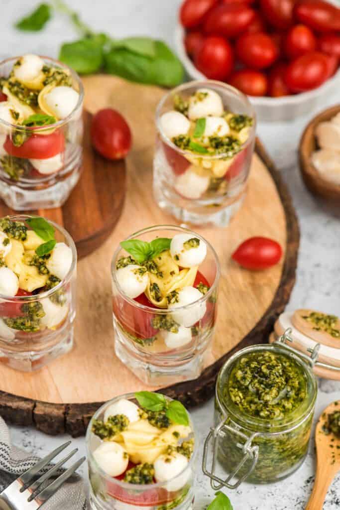 Tortellini Caprese salad cups on a wooden slab surrounded by fresh pesto.