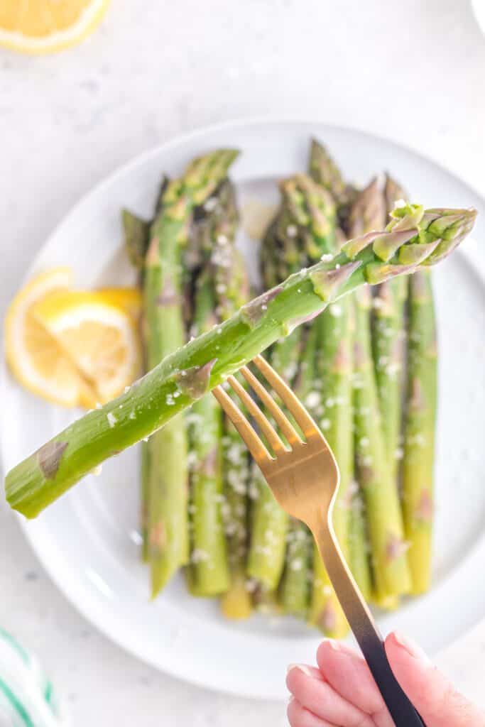 A spear of asparagus on a fork above a platter of cooked asparagus.