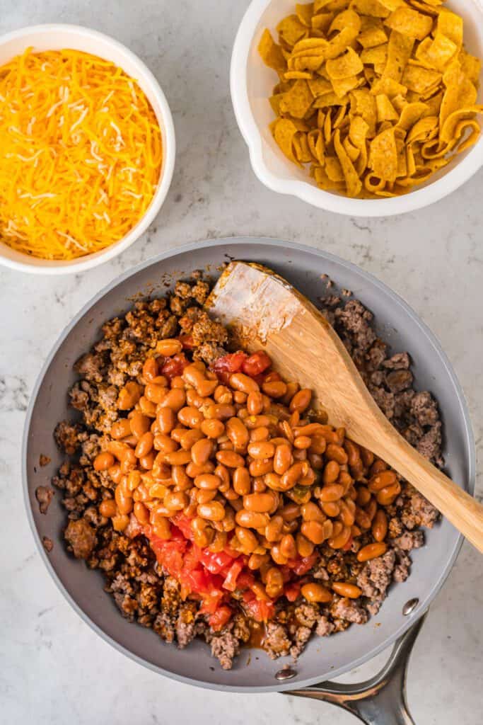 Seasoned ground beef in a skillet topped with tomatoes and beans with a wooden spoon.