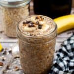 A jar of banana bread overnight oats sitting on a wood countertop with a dish towel.