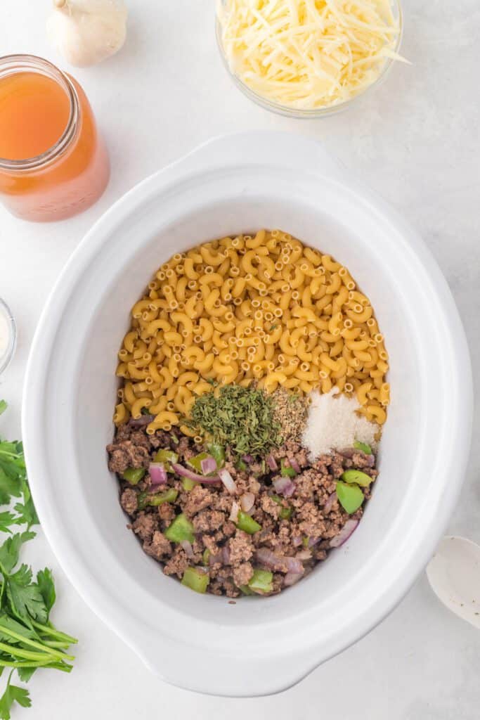 Uncooked macaroni noodles, cooked ground beef, vegetables and spices in a white oval crockpot.
