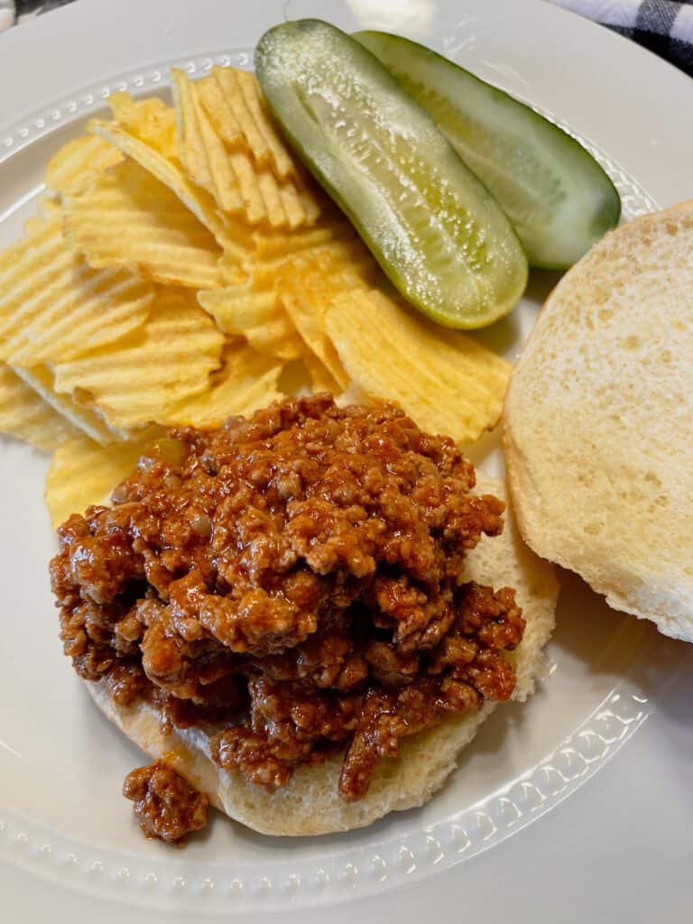Open face sloppy joe spooned on a bun with pickles and potato chips.