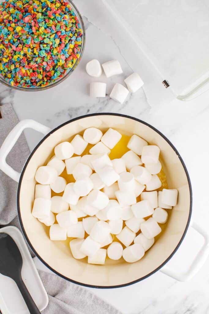 Butter and marshmallows in a large pot with a bowl of fruity pebbles cereal on the side.