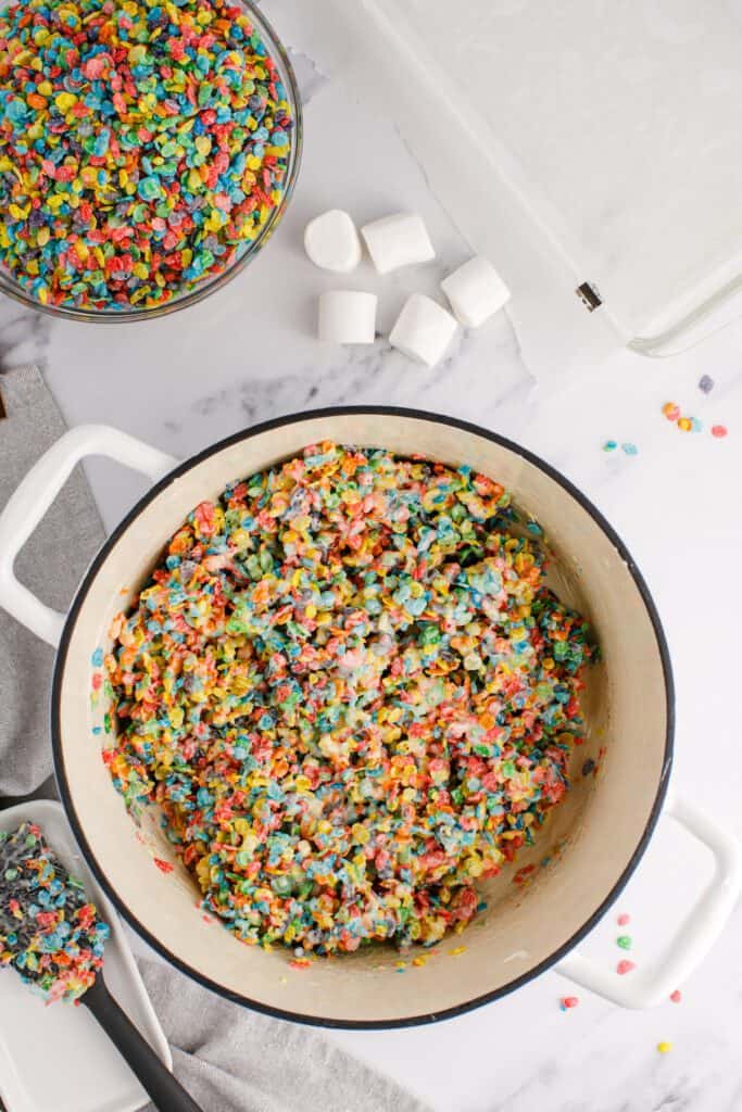 Fruity pebble cereal stirred into melted marshmallows in a large pot.
