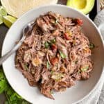 A bowl full of shredded beef with a fork in it surrounded by avocado and flour tortillas.