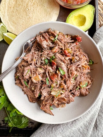 A bowl full of shredded beef with a fork in it surrounded by avocado and flour tortillas.