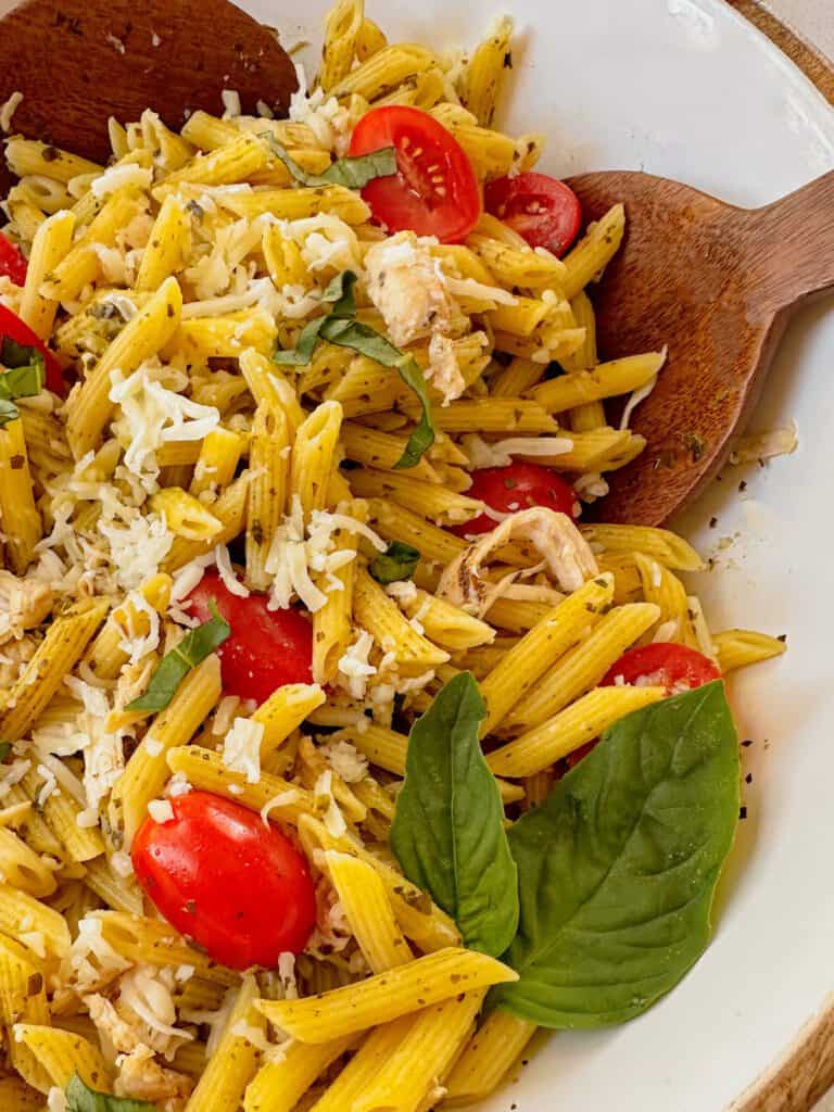 A bowl full of penne pasta, cherry tomatoes, shredded mozzarella and pesto sauce pasta salad with a wooden spoon.