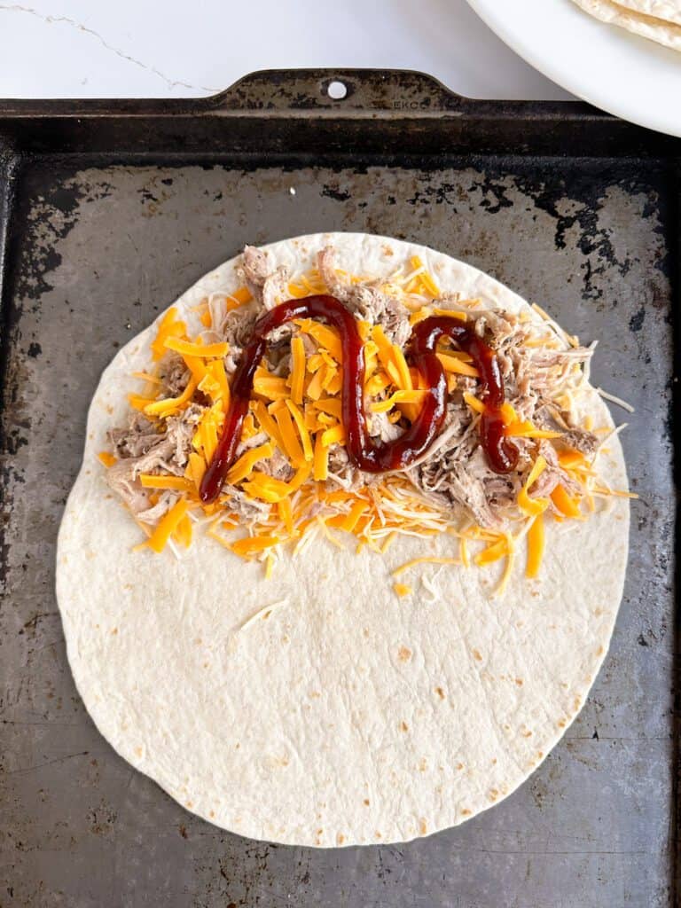 A flour tortilla on a sheet pan topped with shredded cheese, pulled pork and bbq sauce.