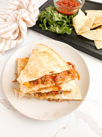 Pulled pork quesadilla wedges on a plate surrounded by chips, salsa and cilantro.