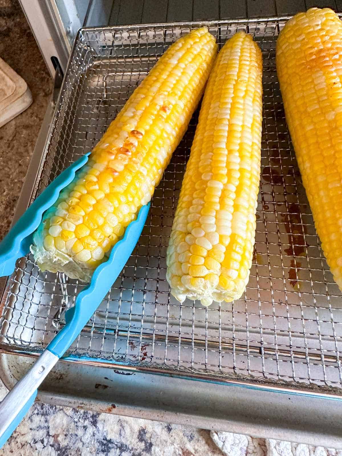 Slightly charred sweet corn in the air fryer being turned over with tongs.