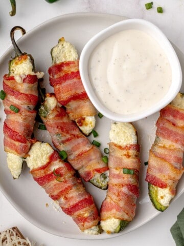 Six bacon wrapped jalapenos on a plate with a bowl of ranch dressing.