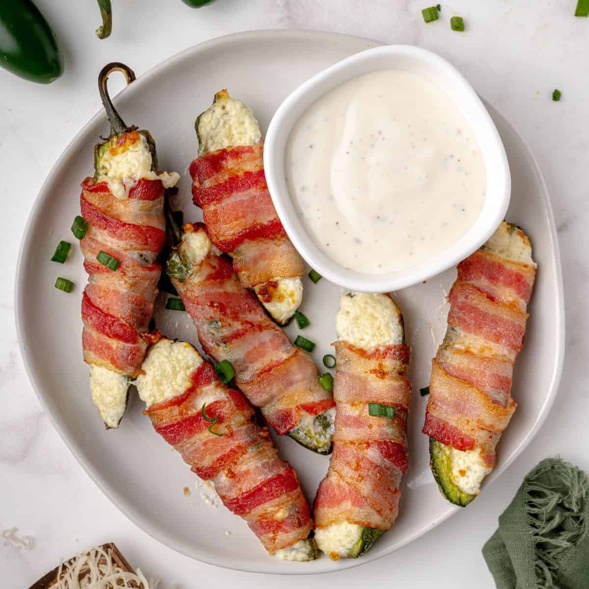 Six bacon wrapped jalapenos on a plate with a bowl of ranch dressing.