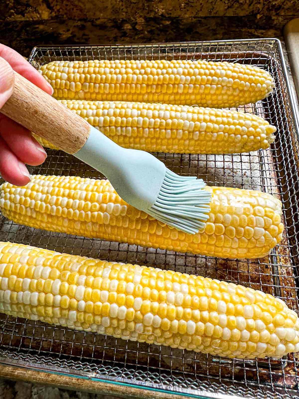 Sweet corn in an air fryer basket being brushed with melted butter.