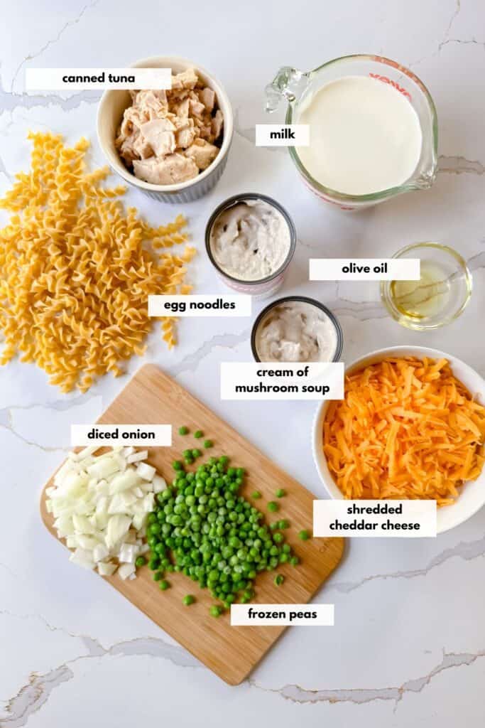 Tuna noodle casserole ingredients including canned tuna, milk, cream of mushroom soup, egg noodles, olive oil, shredded cheese, diced onion and frozen peas.