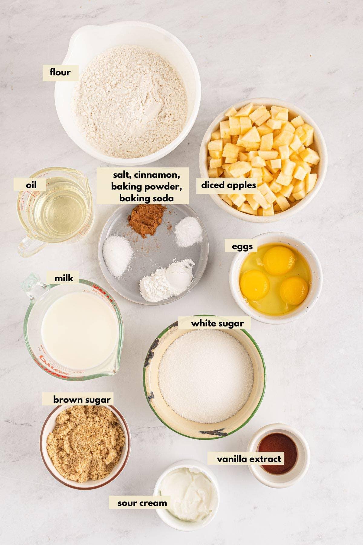Ingredients to make apple muffins including apples, cinnamon, brown sugar, eggs, flour, oil, sour cream.