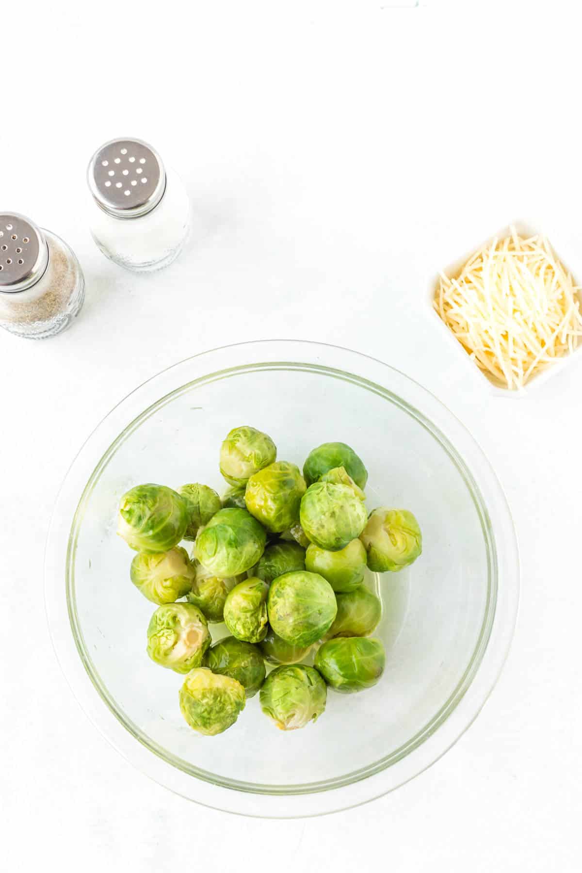 Steamed brussels sprouts in a glass bowl with salt, pepper and grated Parmesan cheese on the side.
