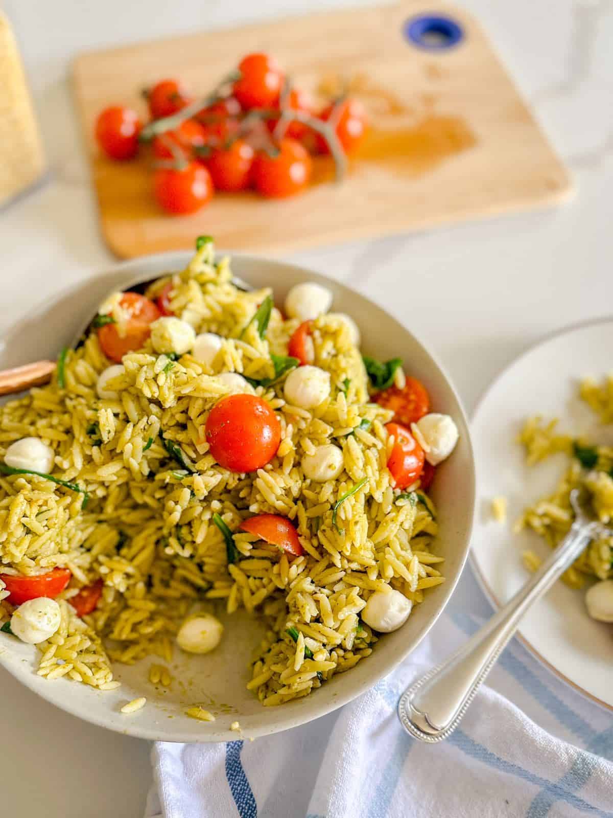 A bowl of pesto orzo salad with cherry tomatoes in the background.