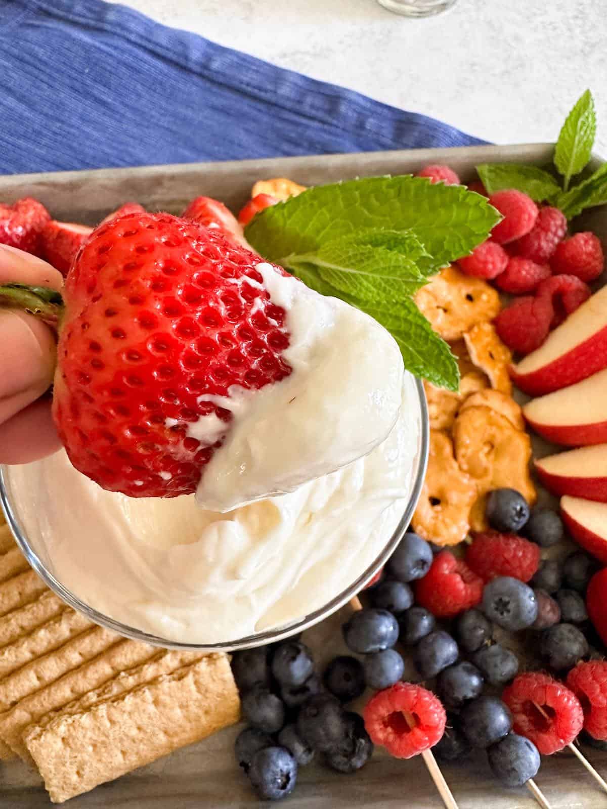 A hand dipping a fresh strawberry into a container of fluff fruit dip.