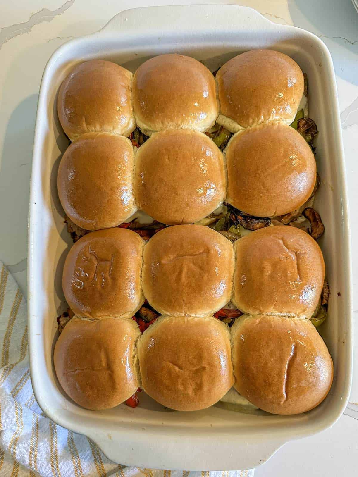Twelve Philly cheesesteak sliders with tops on in a casserole dish.
