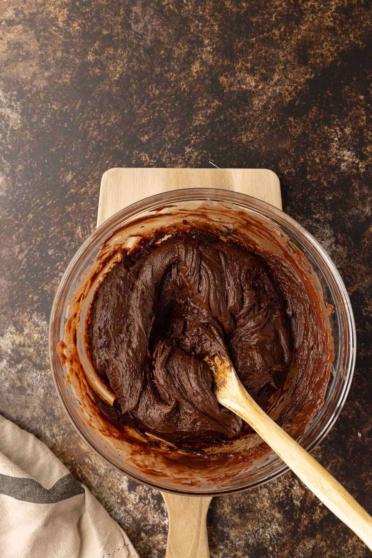 A thick chocolate fudge mixture in a bowl with a wooden spoon.
