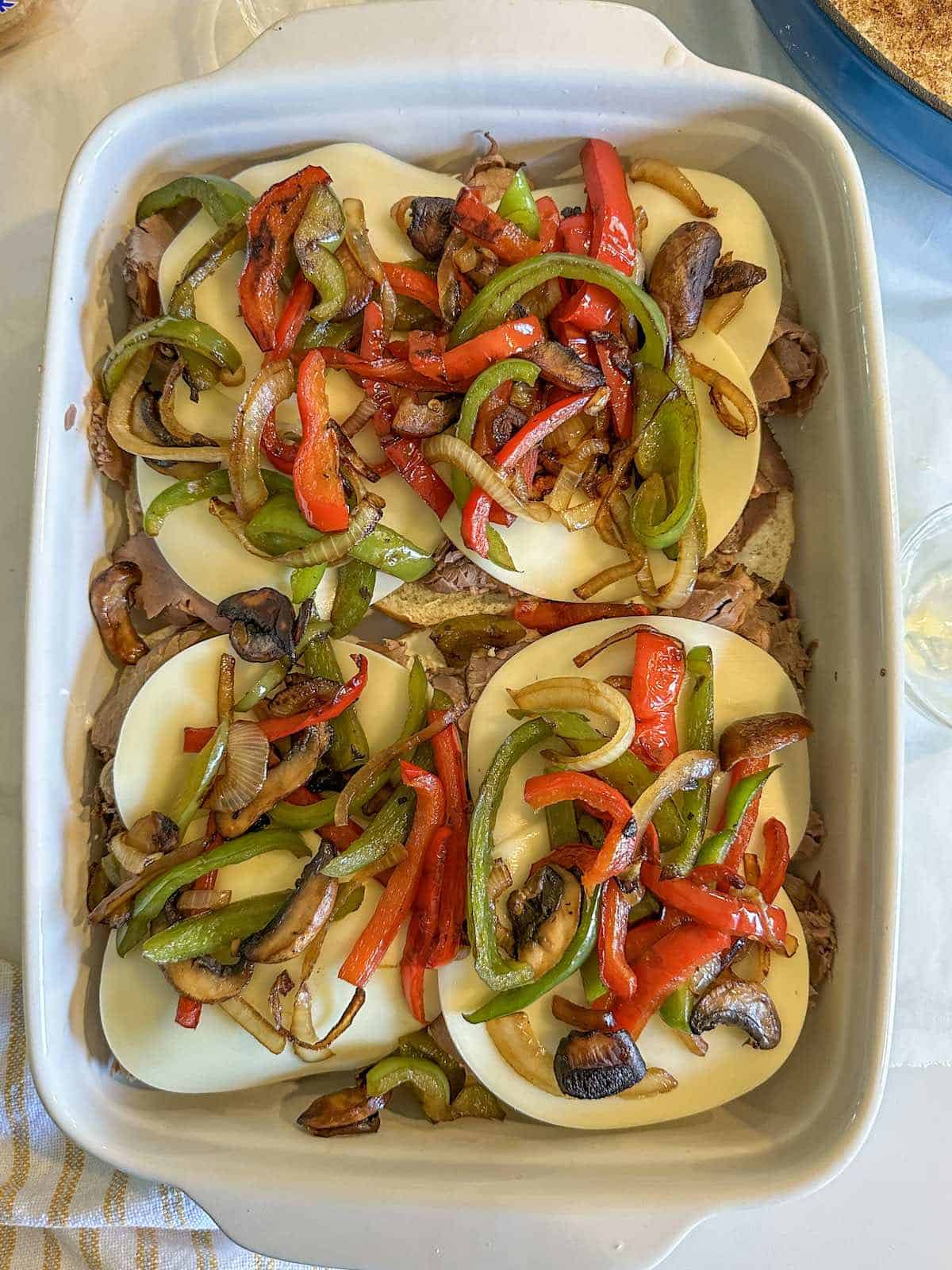 Layered Philly cheesesteak ingredients in a pan with peppers and onions over the top.