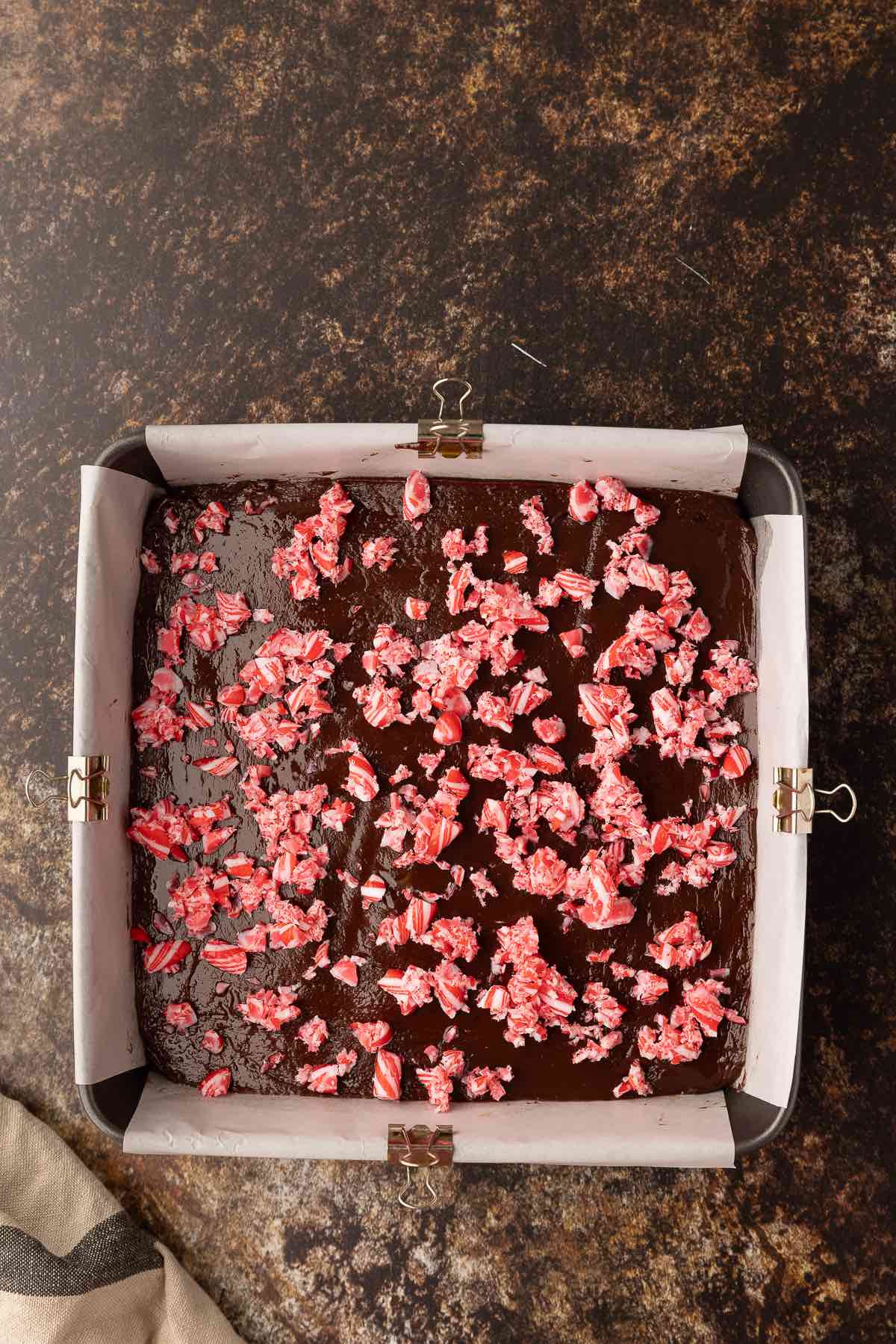 A square pan lined with parchment paper topped with fudge and peppermint candy pieces.