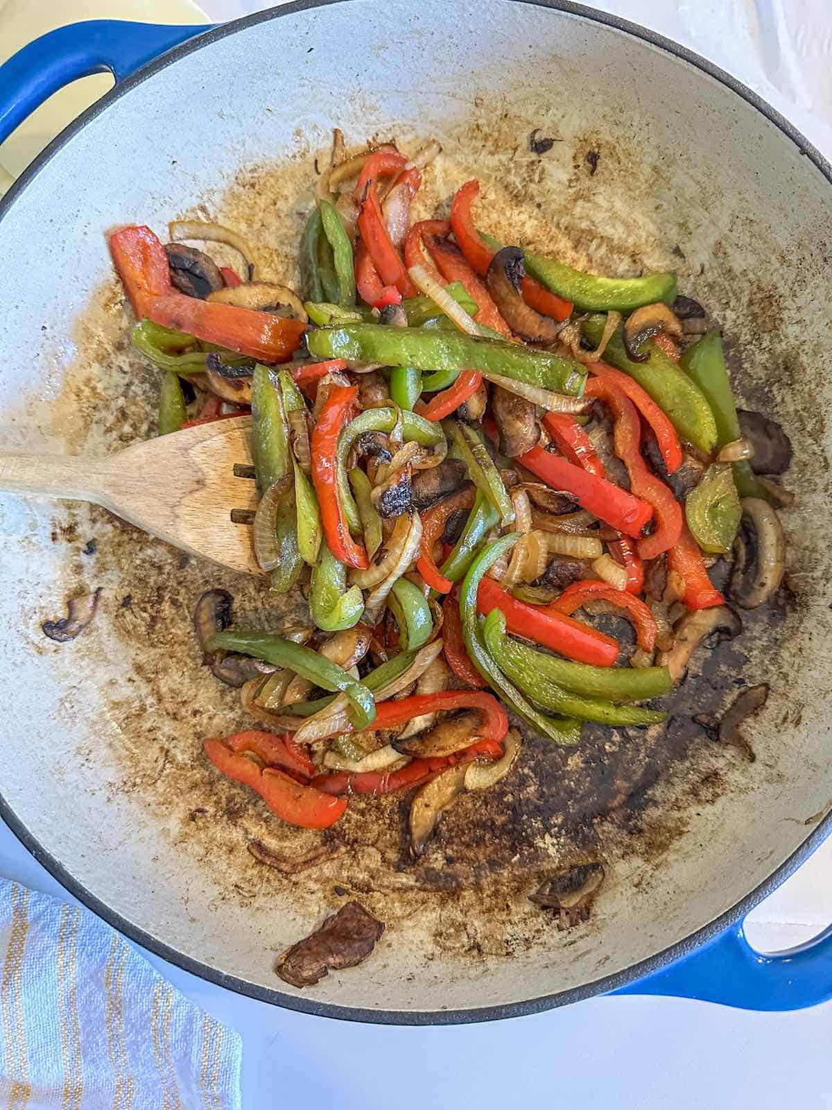 Sauteed green bell pepper, red bell pepper, onions and mushrooms in a skillet with a wooden spoon.