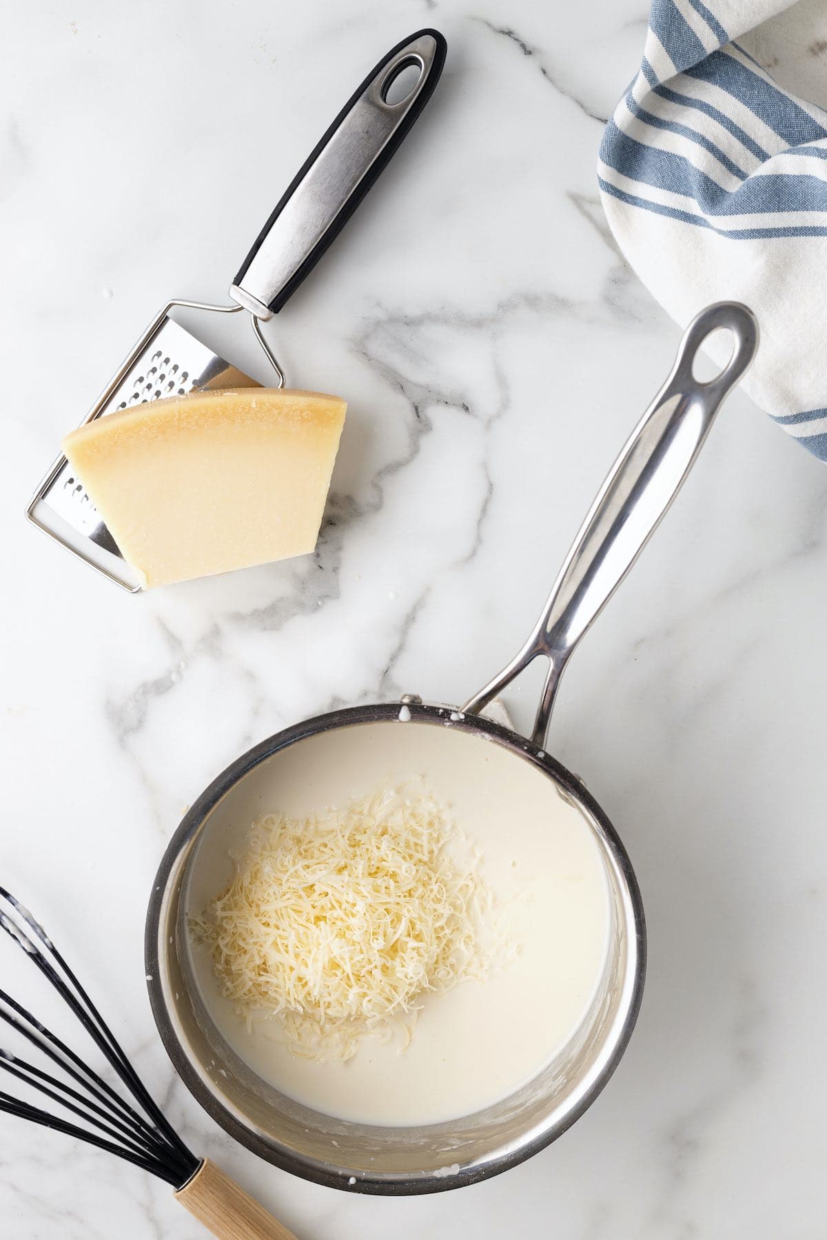 A saucepan with a cream sauce and grated Parmesan cheese on top and a wedge of cheese on the side.