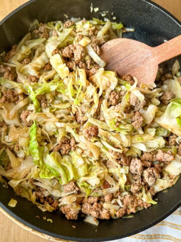 A skillet with ground pork and shredded cabbage and a wooden serving spoon.