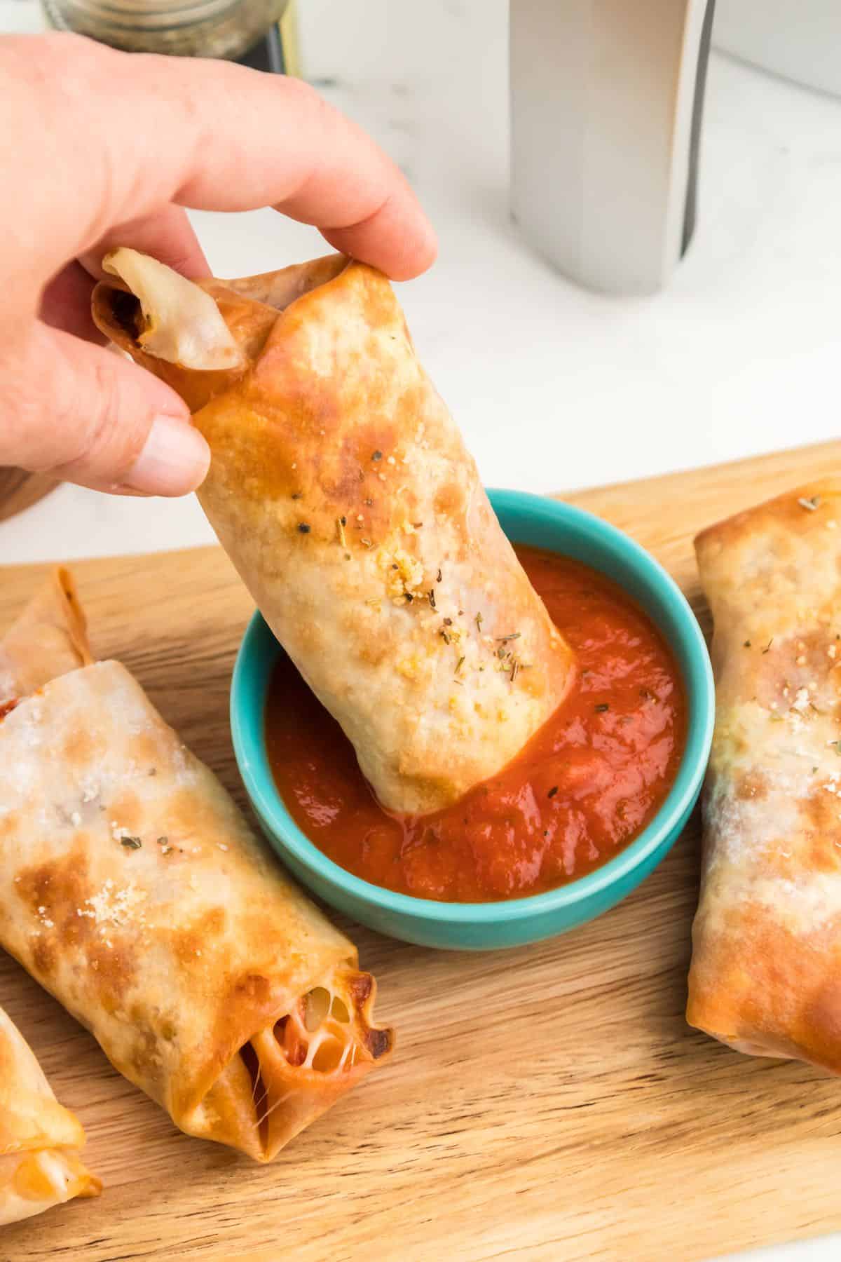 A egg roll with pizza filling dipping into a bowl of marinara sauce.