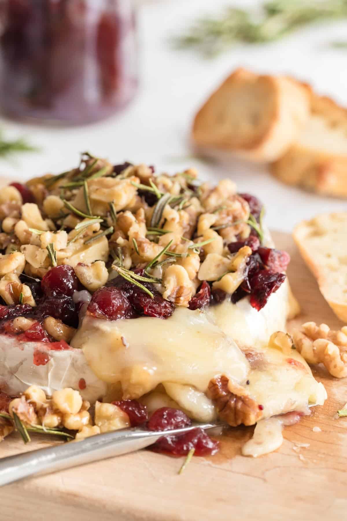 Baked brie with cranberry and walnut topping, surrounded by sliced baguette and crackers, ready to serve at a holiday party.