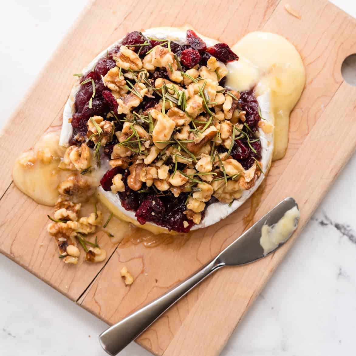 Baked brie with cranberries and walnuts served on a wooden board with crackers and fresh rosemary.