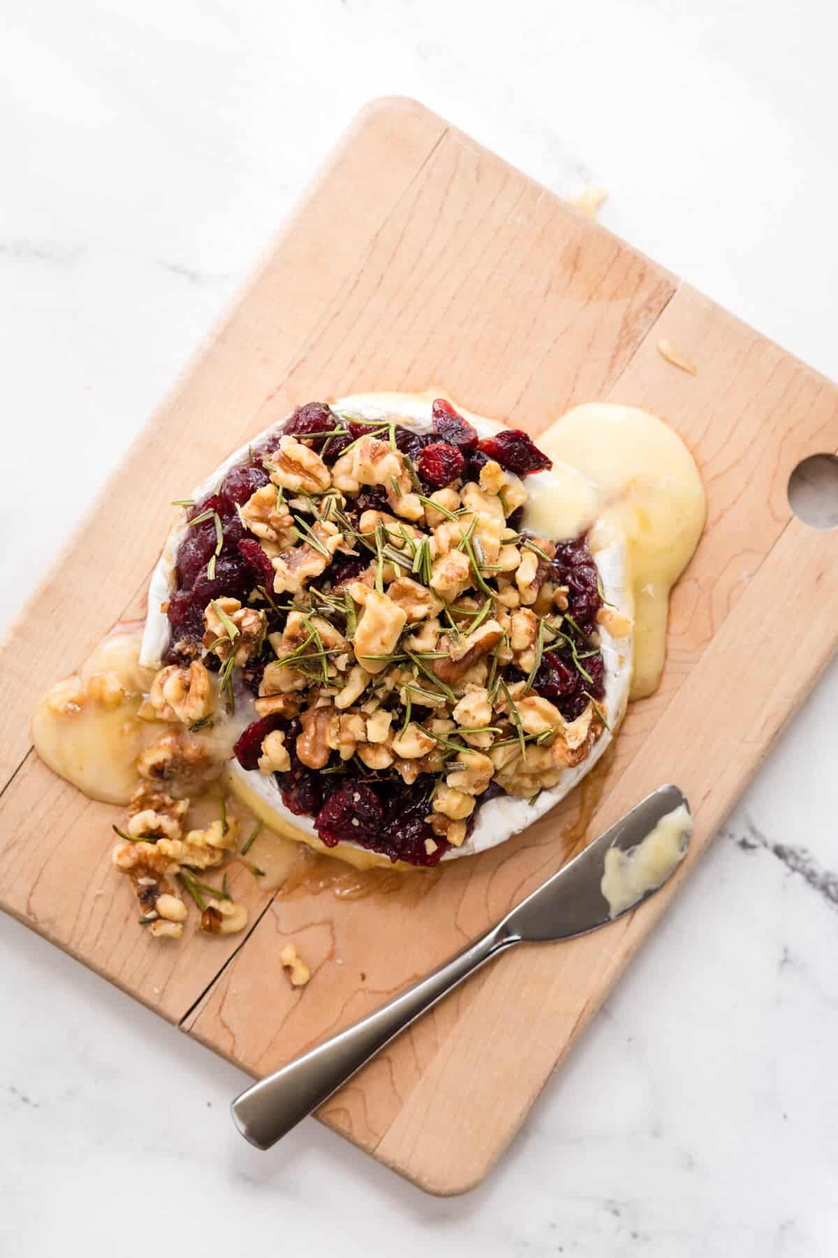 Baked brie with cranberries and walnuts served on a wooden board with fresh rosemary garnish.