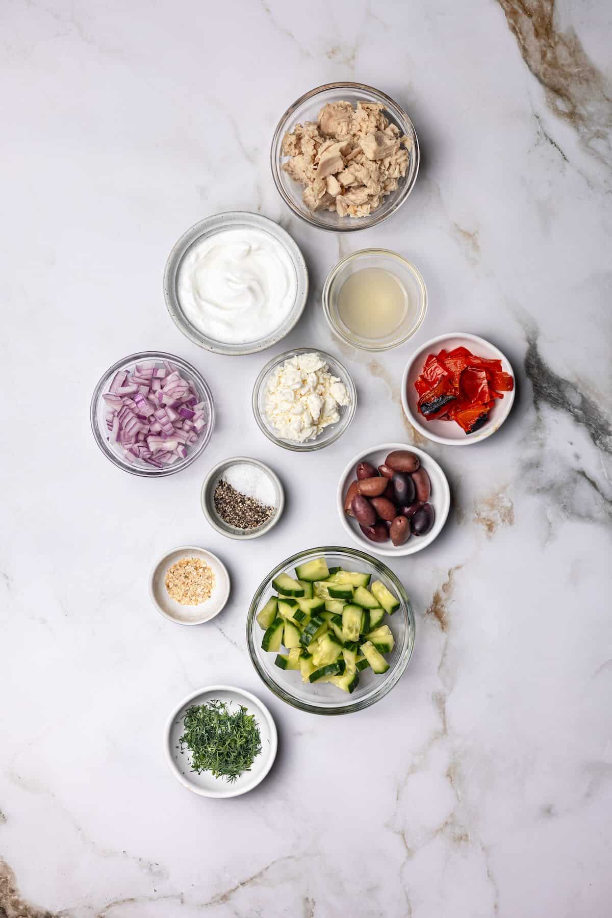 A flat lay of ingredients for Mediterranean tuna salad, including canned tuna, Greek yogurt, lemon, feta, olives, cucumbers, and roasted red peppers.
