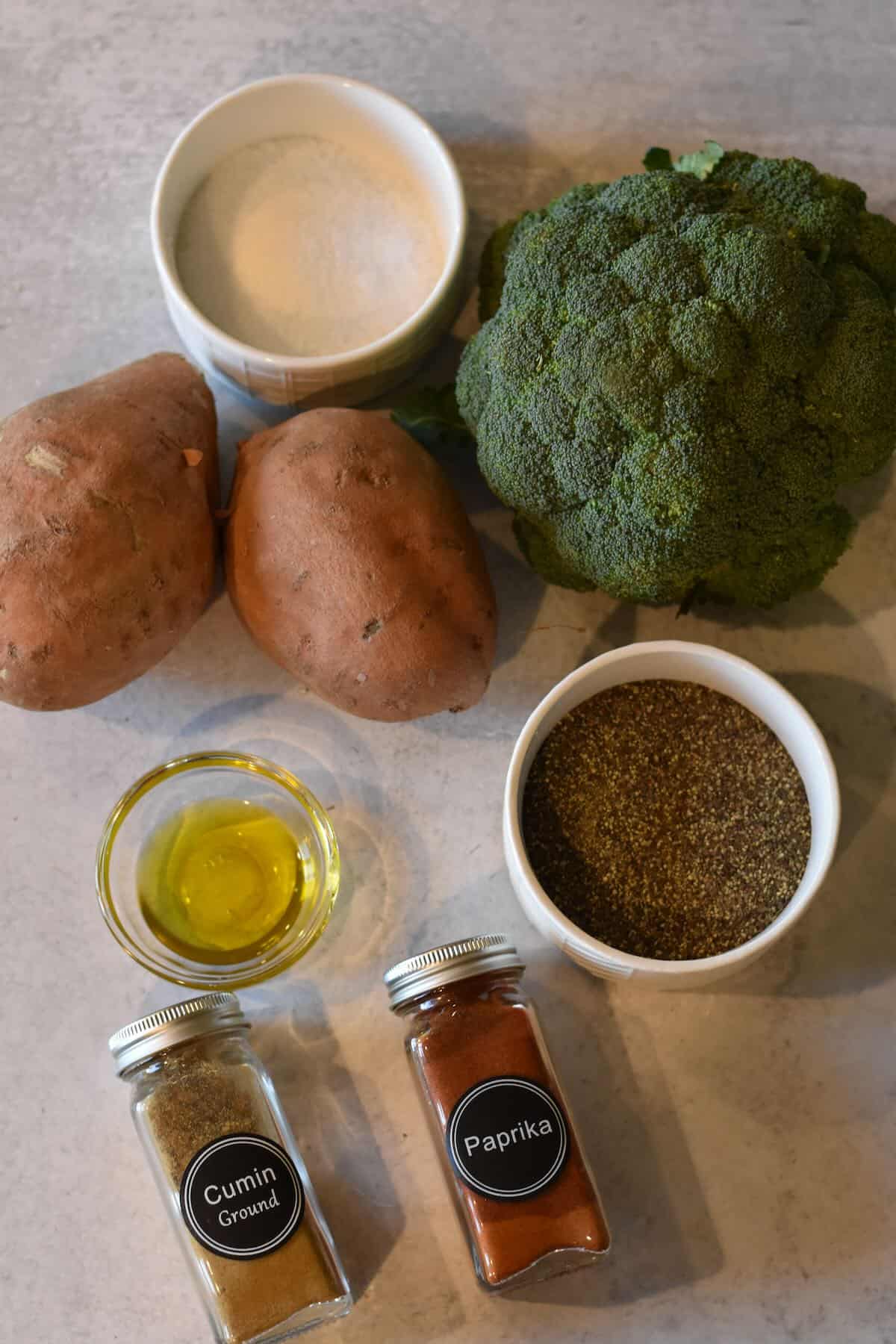 Whole sweet potatoes and broccoli with spices on a countertop.