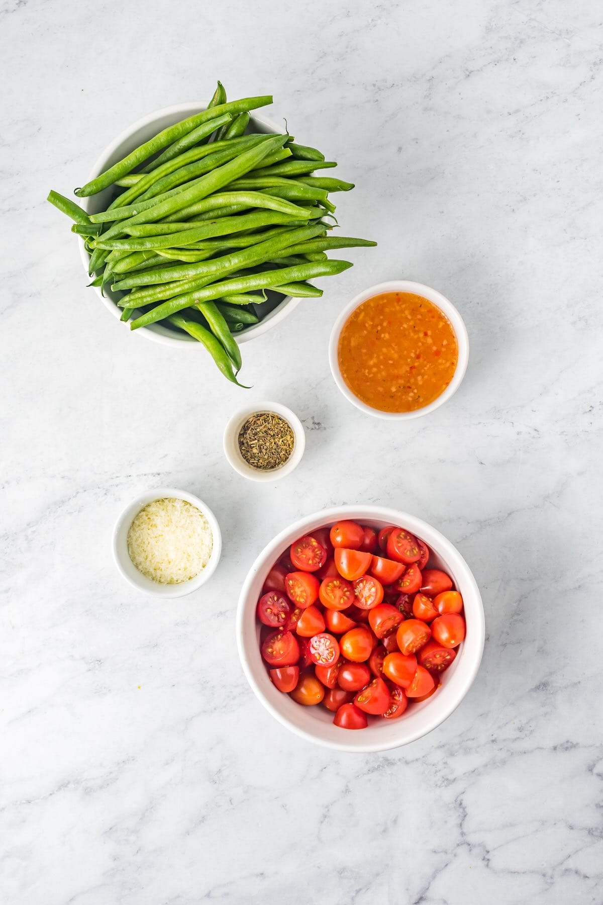 Fresh green beans, grape tomatoes, Italian dressing, Parmesan cheese, and seasonings arranged on a kitchen counter.