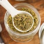 Glass jar of homemade Italian seasoning with spoon for scooping on wood countertop.