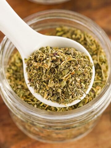 Glass jar of homemade Italian seasoning with spoon for scooping on wood countertop.